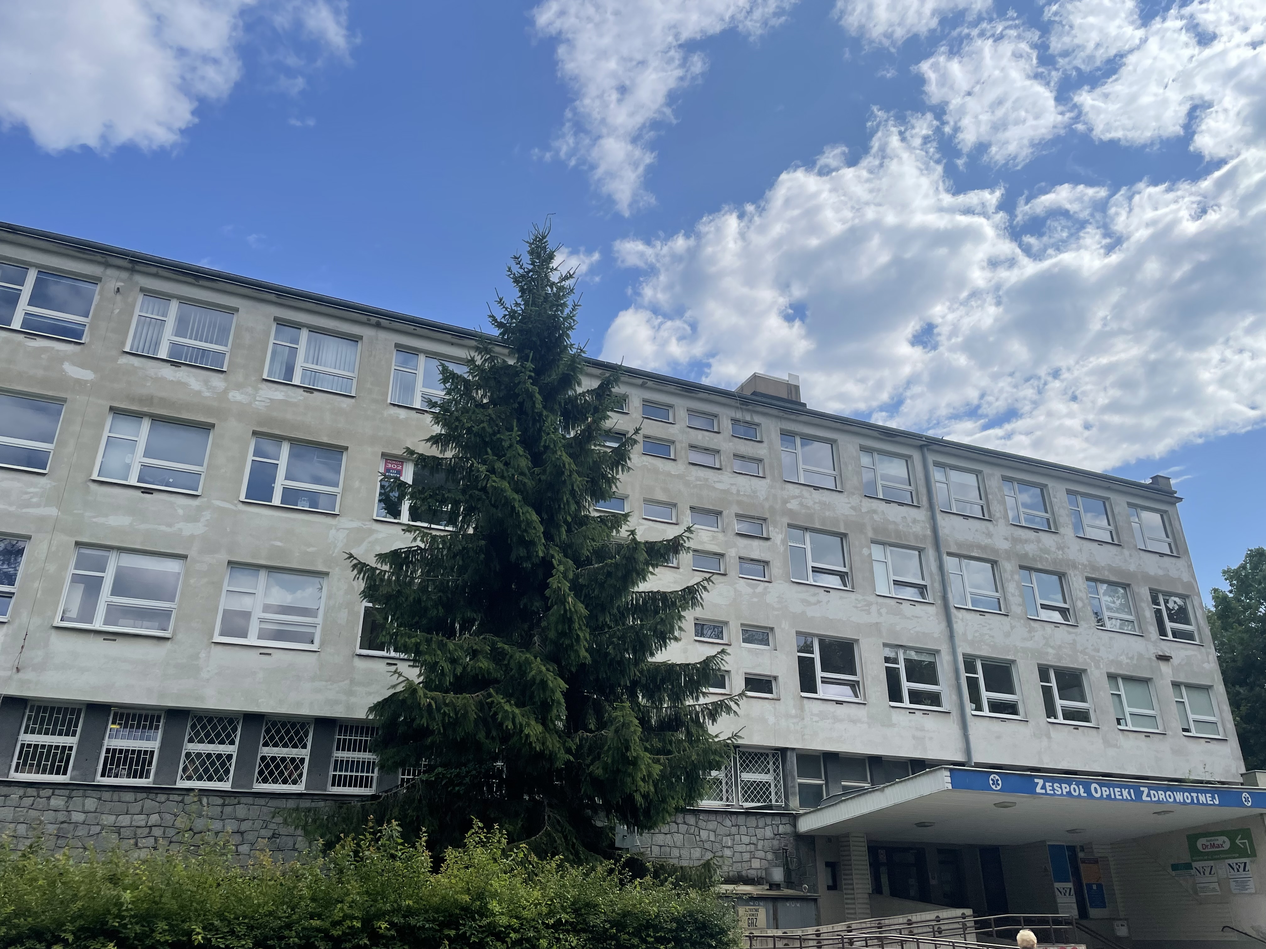 Zdjęcie budynku przy ul. Chrobrego 8/8, w którym znajduje się siedziba spółki. Na zdjęciu widoczne jest wejście do budynku opatrzone napisem Zespół Opieki Zdrowotnej. Widoczny jest również fragment podjazdu dla wózków przy wejściu. z 