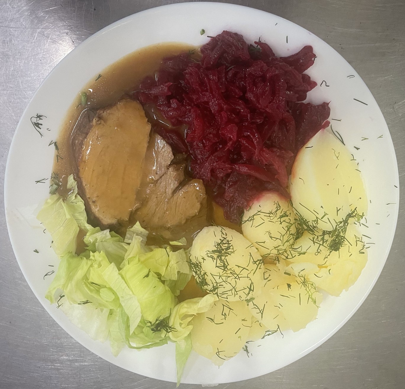 Zdjęcie przykładowego posiłku. Na białym talerzu znajdują się ziemniaki, buraczki, sałata i mięso polane sosem.