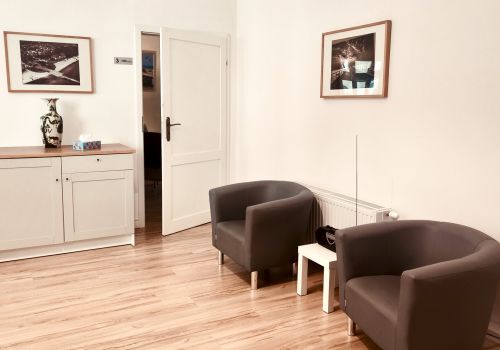 Zdjęcie pomieszczenia terapii grupowej z widocznymi na jasnych ścianach zdjęciami Sopotu, drewnianą podłogą, wygodnymi fotelami i komódką, na której stoją ozdobny wazon i pudełko chusteczek higienicznych