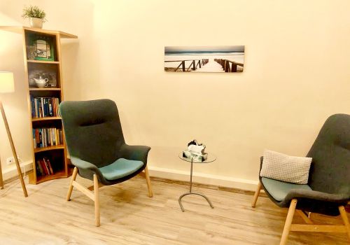 Zdjęcie pomieszczenia terapii indywidualnej. Na zdjęciu widać dwa wygodne fotele, regał z książkami i niewielki stolik, na którym umieszczono pudełko z chusteczkami. Na ścianie wisi obraz prezentujący sopockie molo.