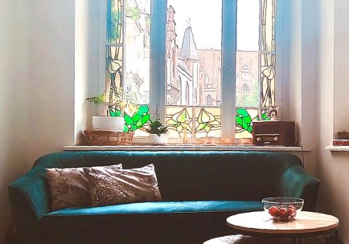 Kolejne zdjęcie werandy - widiczne jest na nim całe okno z witrażem oraz kanapa i niewielki, podwójny, stolik kawowy, na którym stoi misa z owocami..